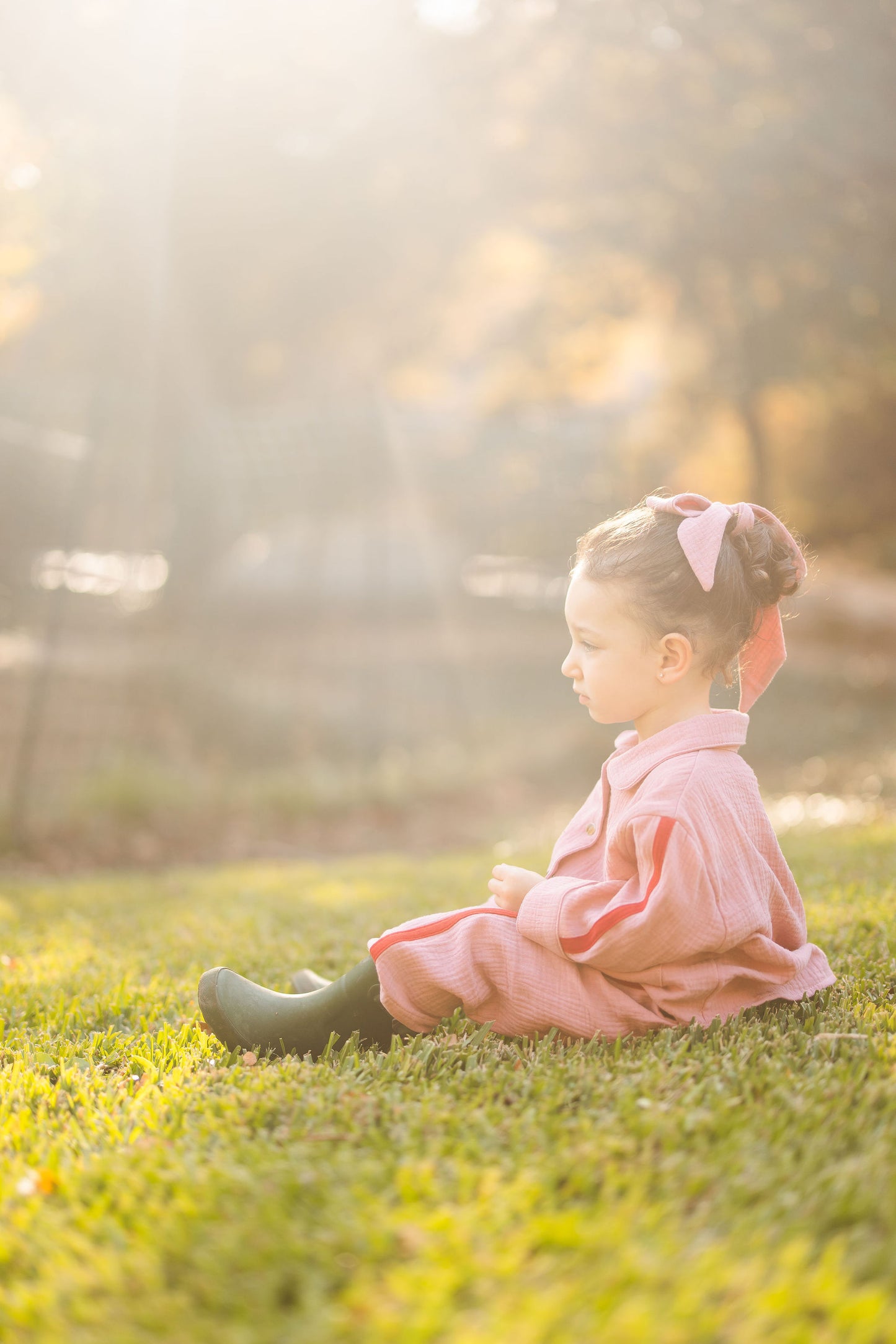 Child in a pink lounge wear set sitting on grass with a blurred natural background